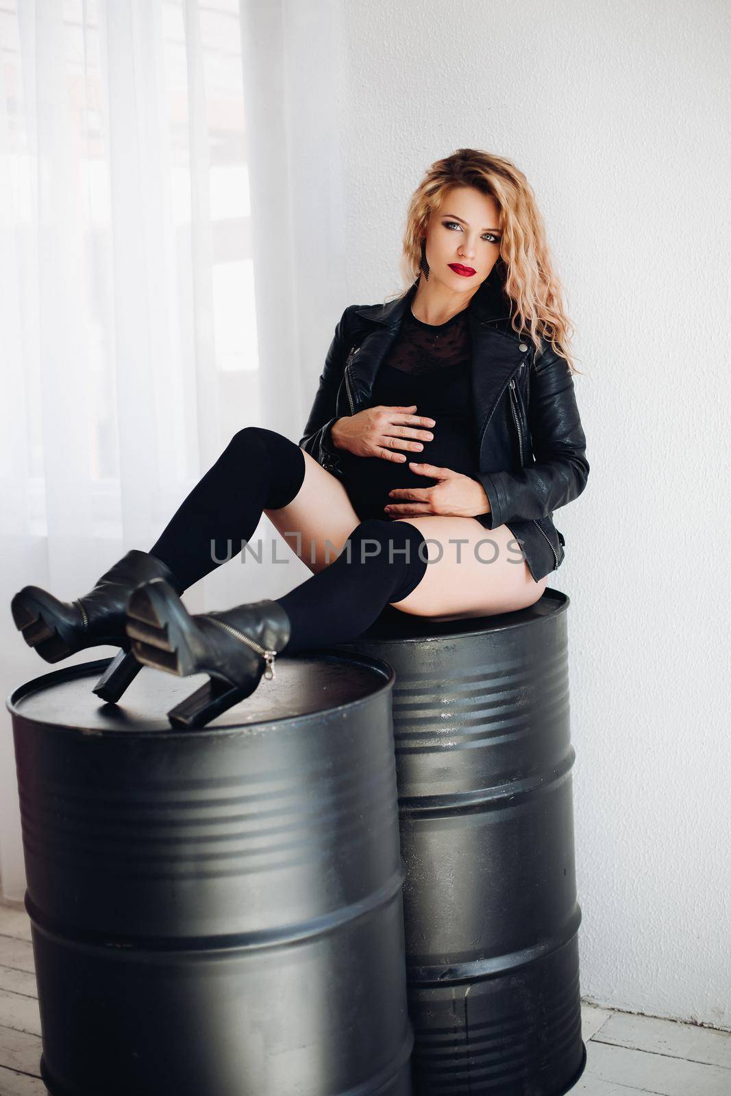 Studio portrait of pregnant lady in stylish leather jacket, knee socks and high heels posing on black containers embracing belly and looking at camera.