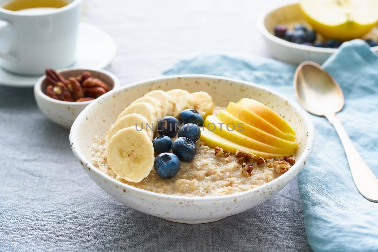 Whole oatmeal, large bowl of porridge with banana, blueberries, apple, nuts for breakfast, morning meal. Side view, close up, gray table. Vegan tasty healthy diet.