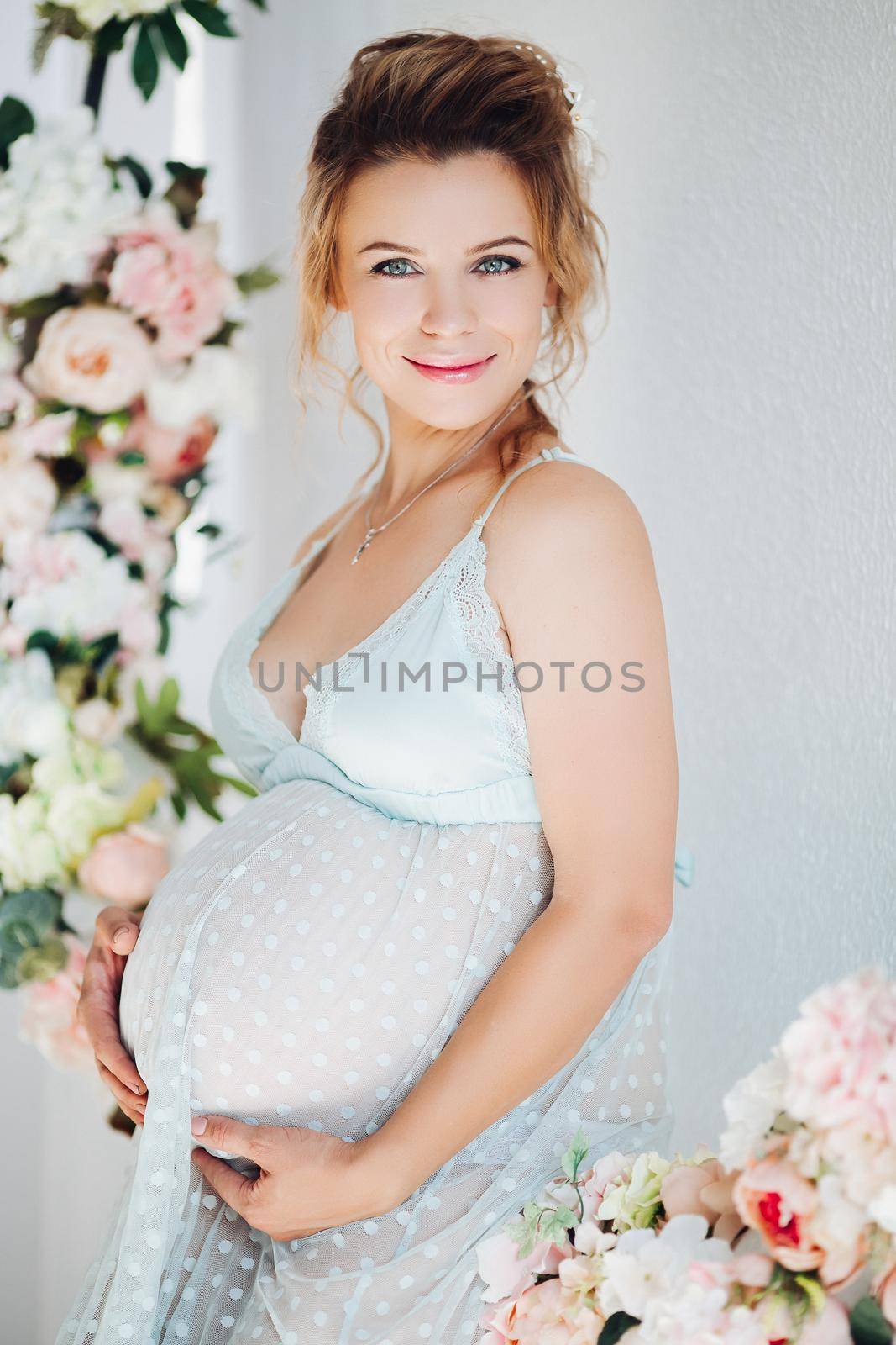 Beautiful and sensuality pregnant woman with blue eyes and hairstyle, wearing in lingerie dress. Smiling young mom expacting baby, embrasing stomach and smiling at camera. Flowers on background.