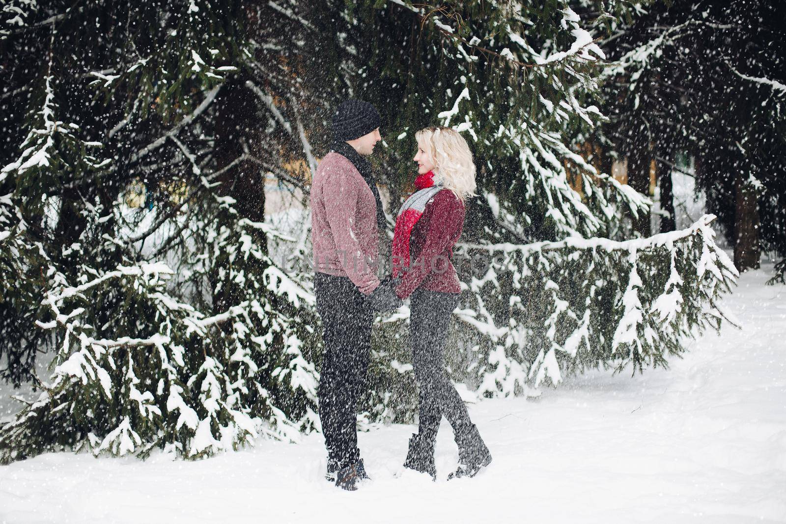 Young couple standing and looking each other in snowing forest. Blonde wife and her husband wearing red sweaters. Woman with volumed hair touching her significant other. Concept of tendernness.