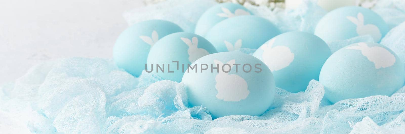 Easter banner. Holiday. Light white background, gentle pastel colors. Blue eggs with image of rabbit in basket. Flowers in background