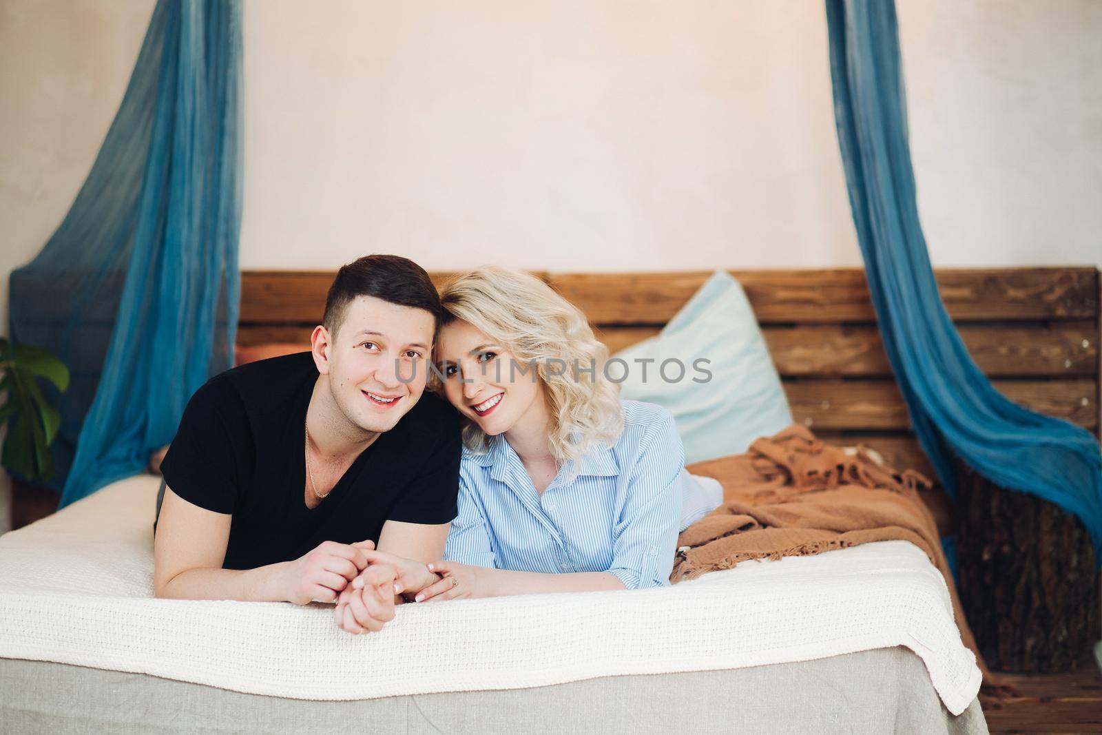 Young couple laying together on belly in white bed. Smiling and holding hands of each other, looking at camera. Blonde woman in striped shirt and a man in black tshirt.Concept of love and relationship.