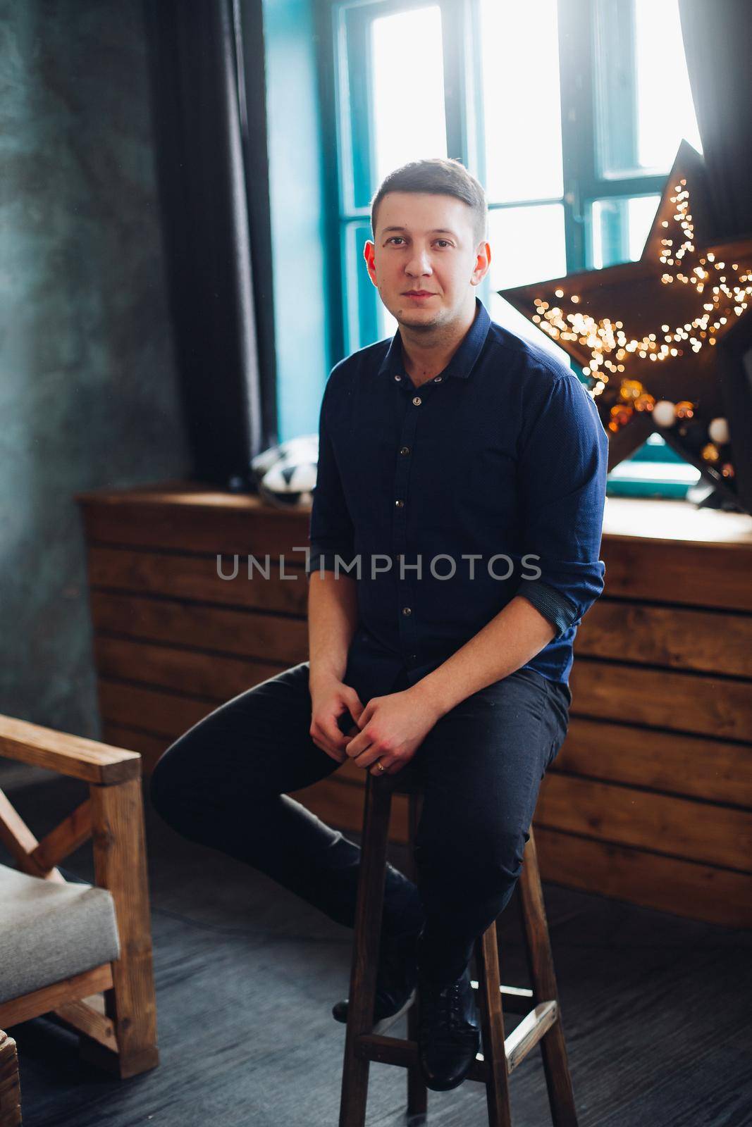 Portrait of smiling handsome man sitting on chair in decorated studio, looking at camera. Confident brunet male in black wear against ligth stars on windowshill.