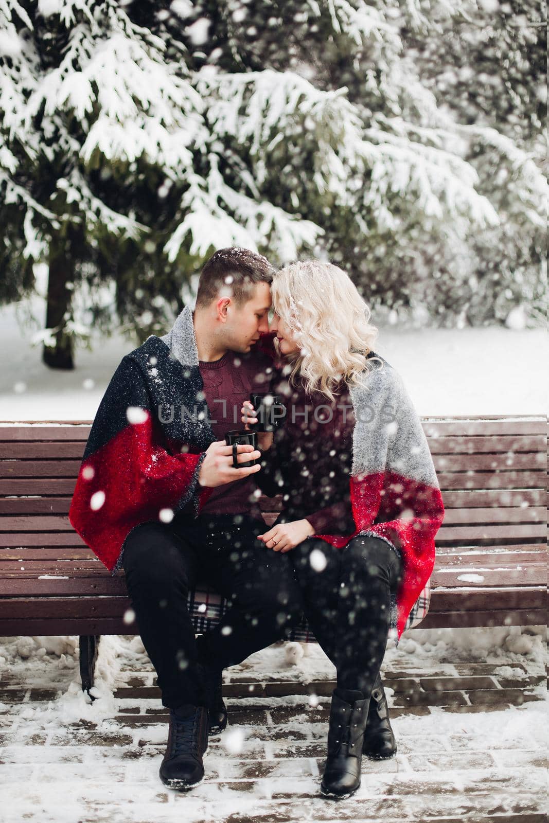 Amorous couple boyfriend and girlfriend posing and sitting on bench in winter forest. Pretty blonde and smiling man looking at camera and holding cups with coffee in hands. Concept of love.