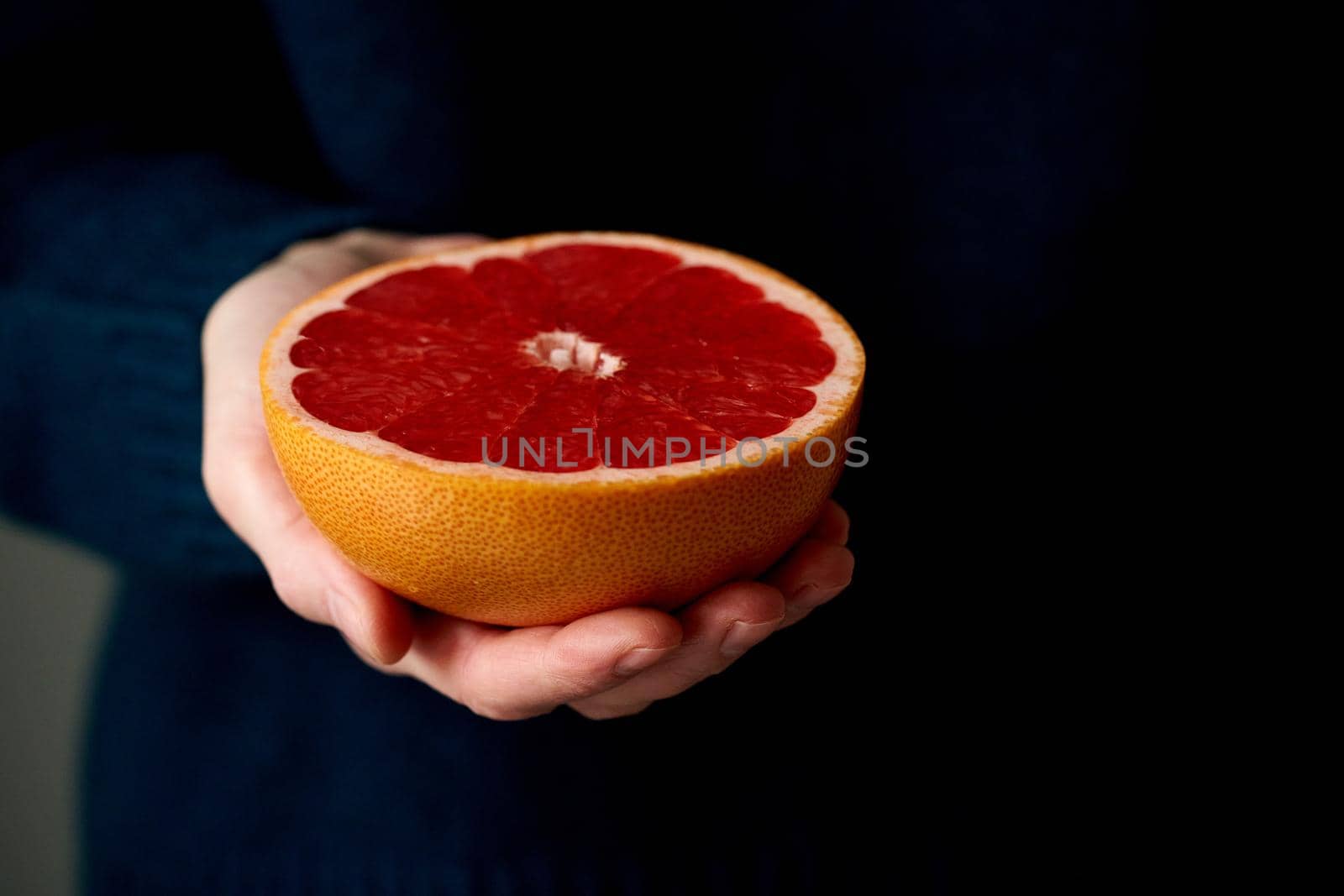 Close-up of woman's hand holding fresh halved red bright citrus fruit grapefruit on dark background. Lifestyle, natural light from window. Vitamins