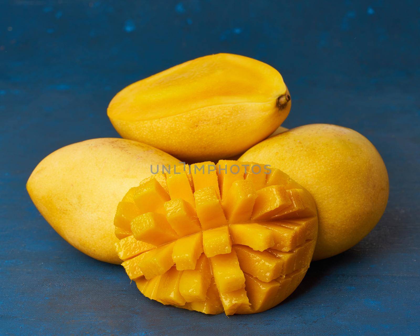 Four whole mango fruits on dark blue table and cut into slices. Large juicy bright ripe yellow fruits on color background, close up, copy space