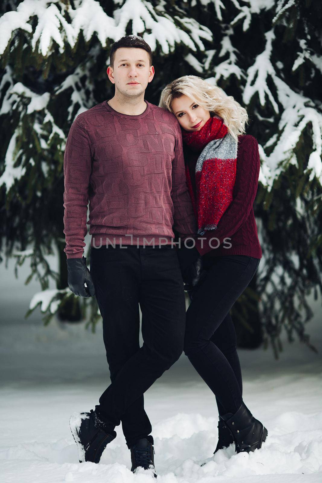 Close up of blonde wife leaning on shoulder her husband and smiling at camera. Beautiful couple of girlfriend and boyfriend standing in snowing forest and posing together.Concept of Christmas holiday.