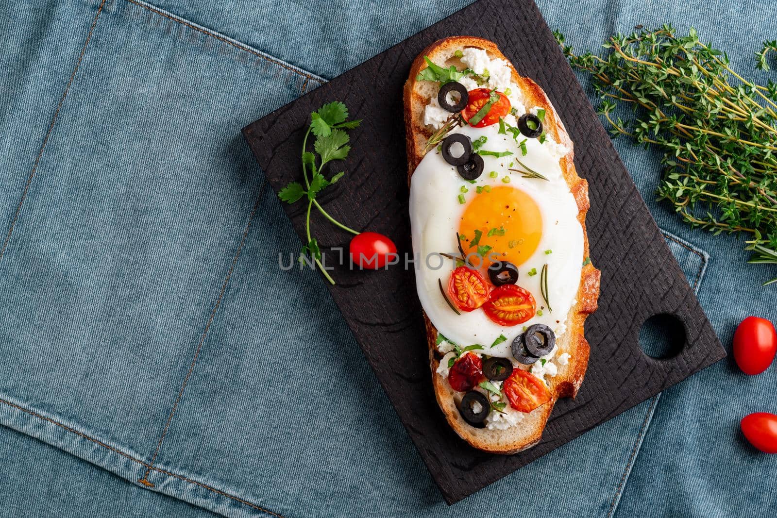 Toasted bread toast with fried eggs with yellow yolk and tomatoes, olives, sprinkled with herbs on dark wooden serving board on blue denim napkin, top view, copy space