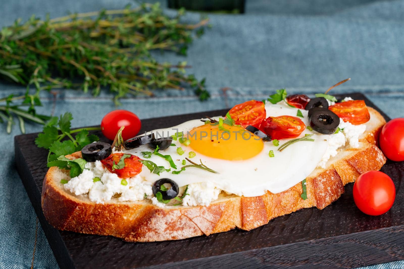 Toasted bread toast with fried eggs with yellow yolk and tomatoes, olives, sprinkled with herbs on dark wooden serving board on blue denim napkin, side view