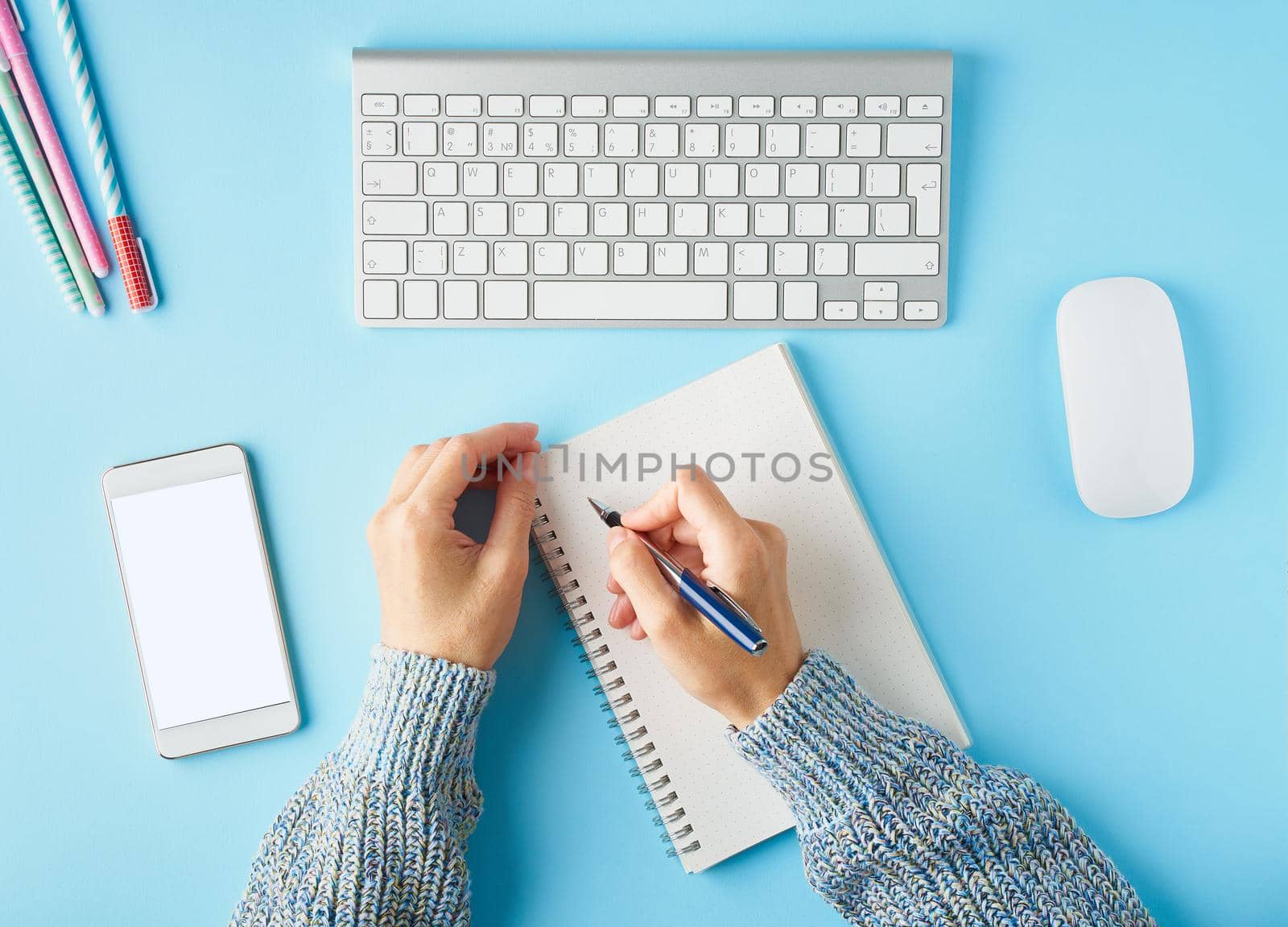 Faceless woman writing in notepad. Mobile phone with blank white screen display. Bright blue modern desk with smartphone. Top view. Copy space, mock up.