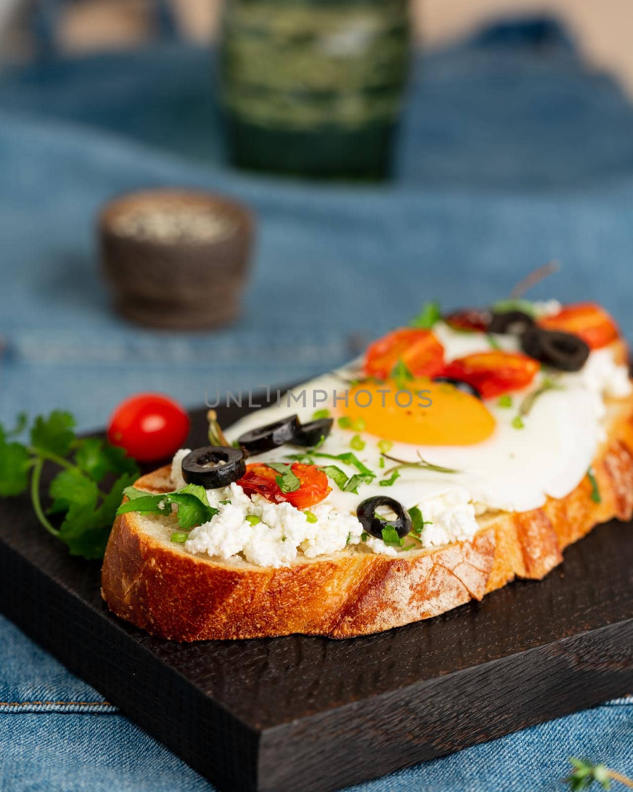 Toasted bread toast with fried eggs with yellow yolk and tomatoes, olives, sprinkled with herbs on dark wooden serving board on blue denim napkin, vertical