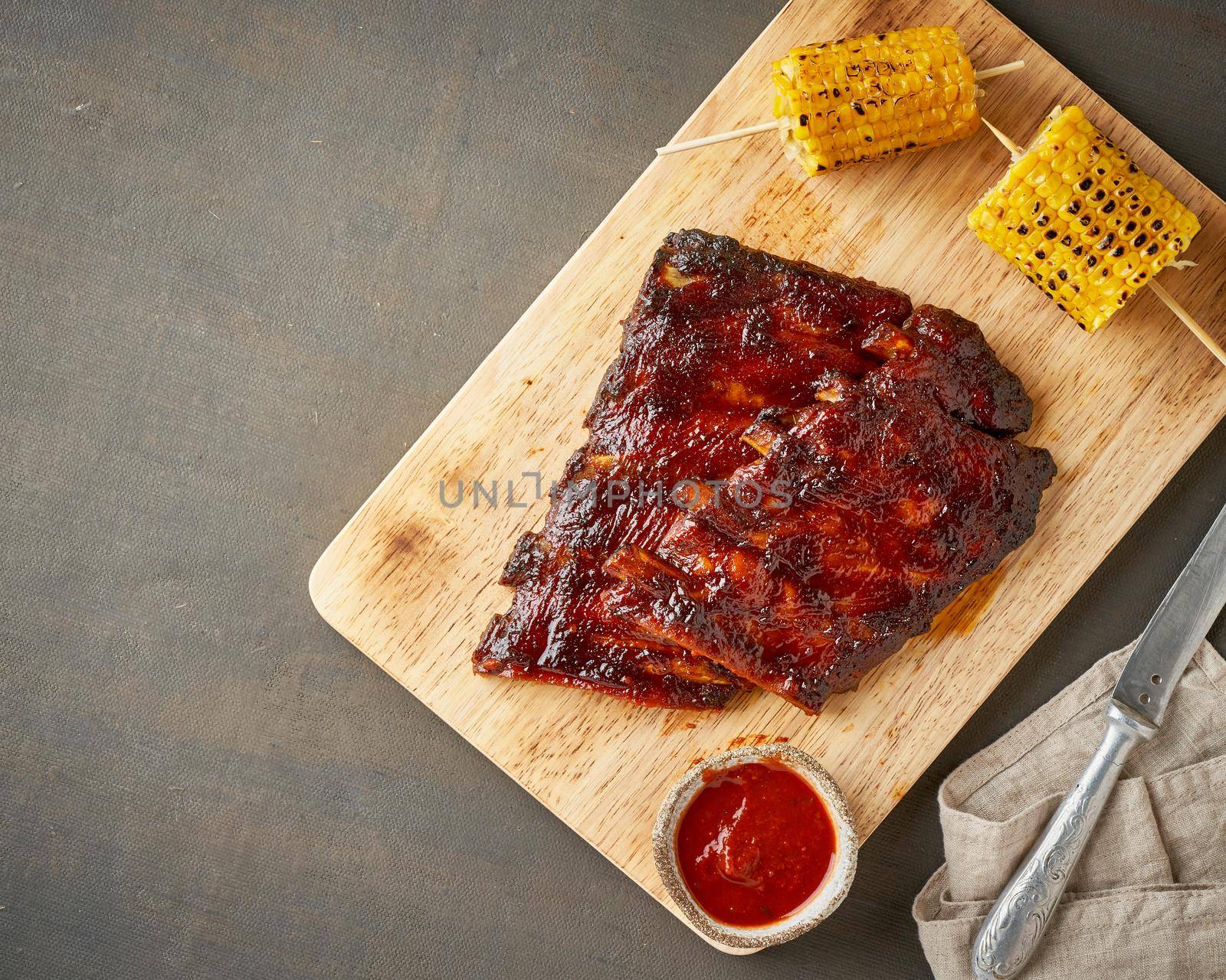 Barbecue pork ribs. Slow cooking recipe. Whole pickled roasted pork meat with red sauce and corn on cutting board, copy space, top view. Brown background
