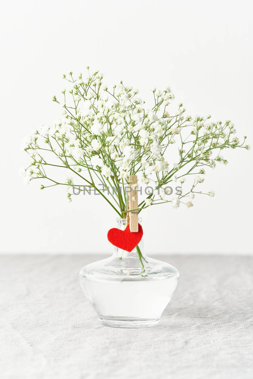 Valentine's Day. Delicate white flowers in glass vase. Red felt heart - symbol of lovers. Light white gentle pastel background. Scandinavian minimalism. Side view, copy space, vertical