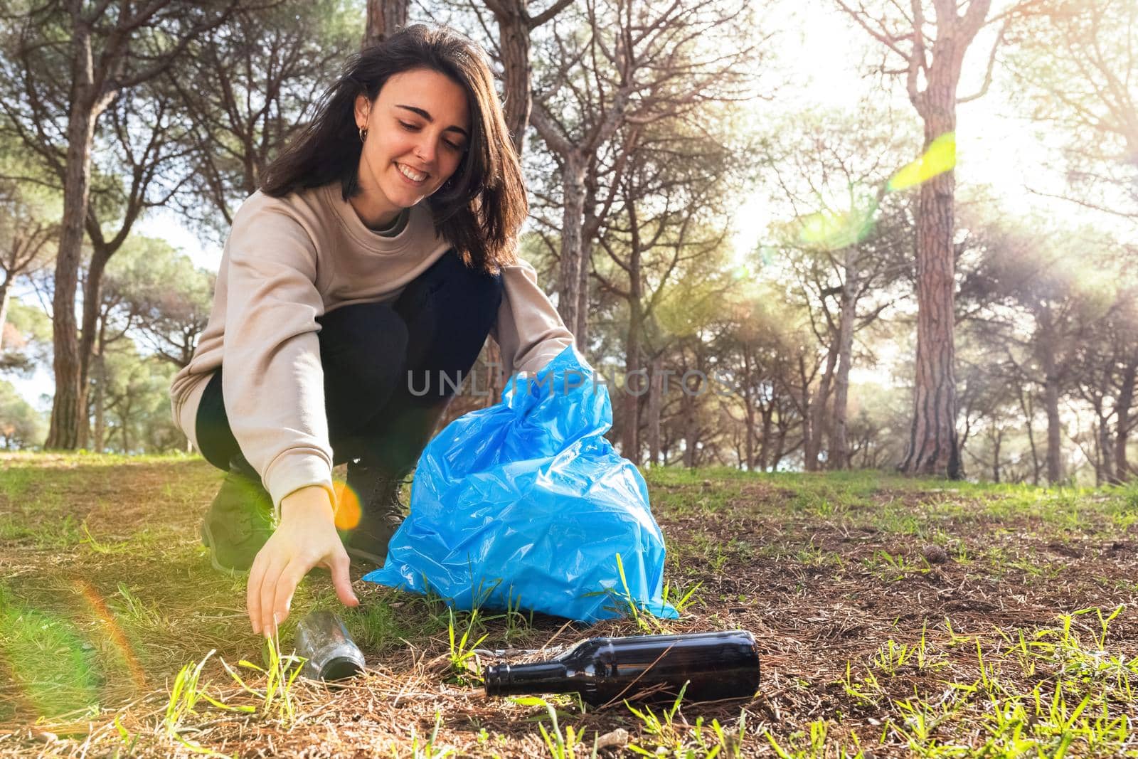 Smiling young caucasian woman pick up plastic bottle in the forest. Copy space. Environmental activist concept.