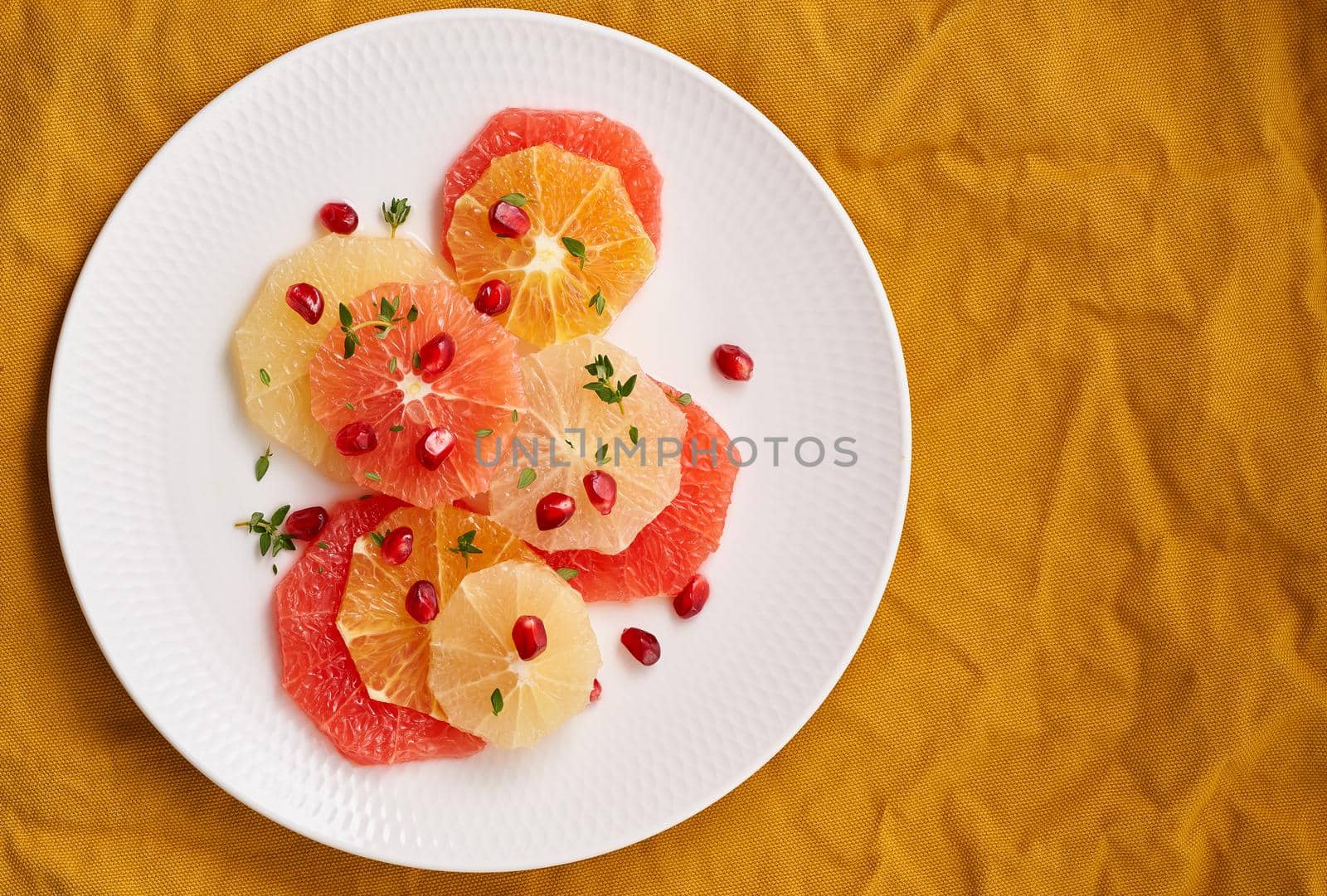 Bright mix of citrus fruits. Salad of mix sliced round slices of red and white grapefruit sprinkled with pomegranate seeds on white plate on yellow linen. Copy space