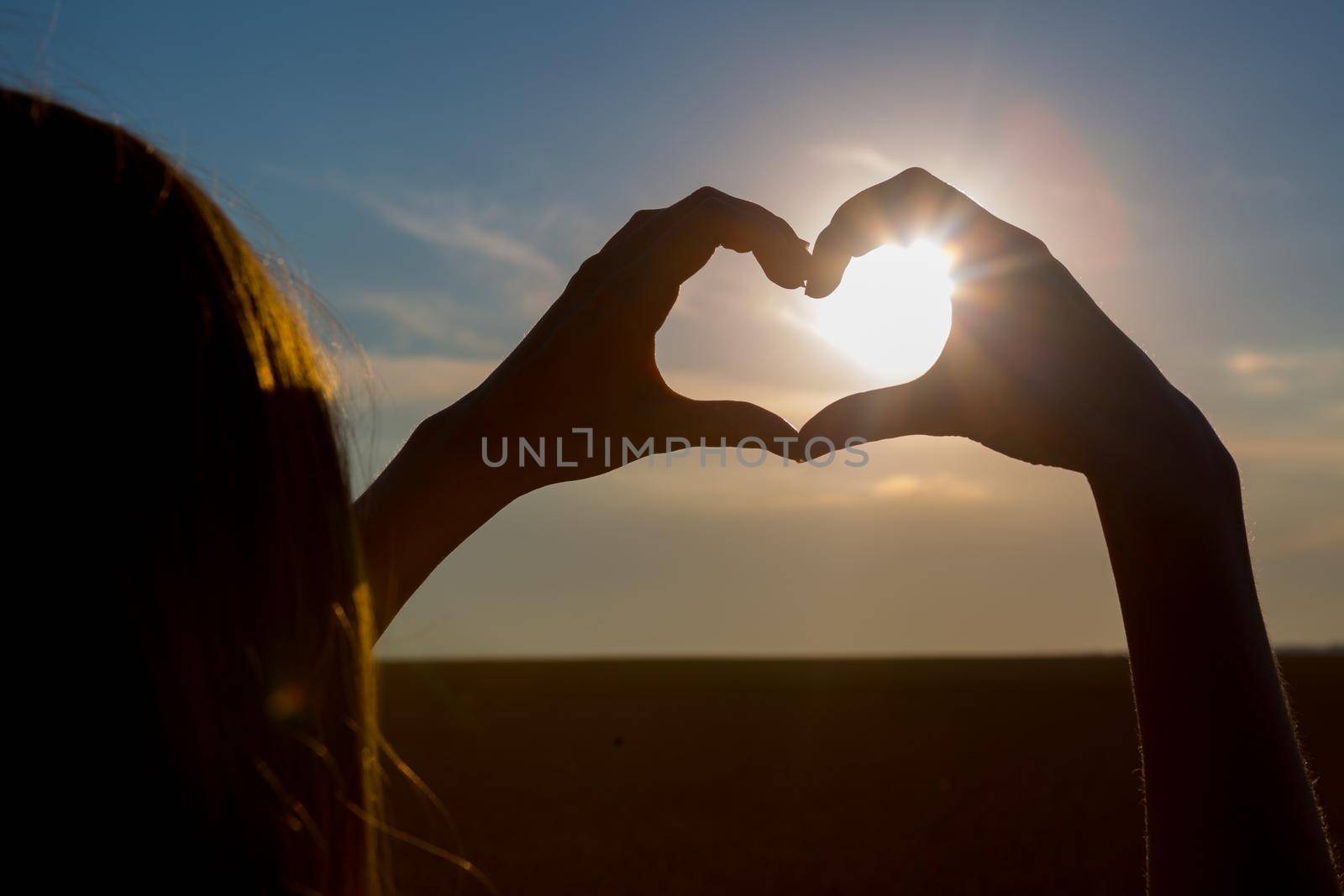 Girl making heart symbol with her hands at sunset on nature background.