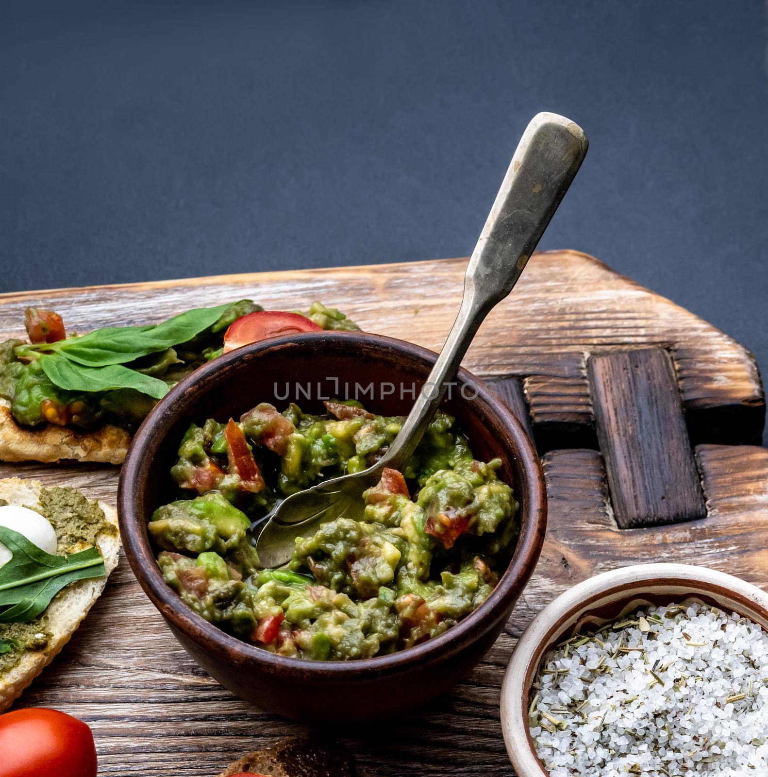 Traditional mexican guacamole and salt with herbs on wooden board with bruschettas closeup. Toasted bread with pesto, basil and tomatoes served with avocado sauce
