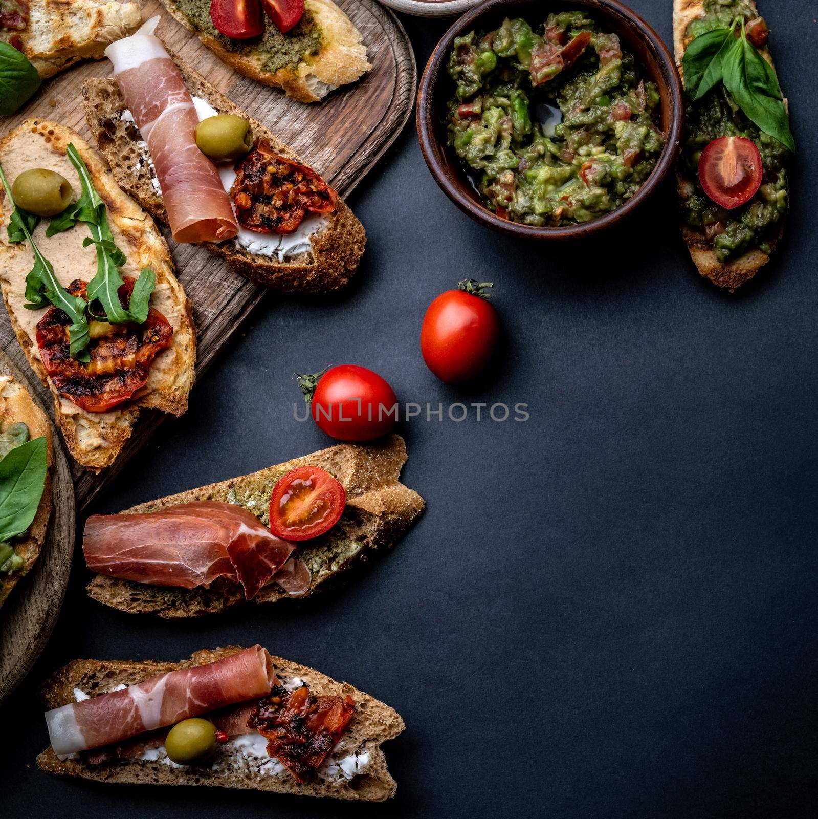 Set of bruschettas with jamon, olives, pesto, tomatoes, basil and mozzarella served on wooden board with guacamole and cherries. Traditional mediterranean antipasti food composition with copy space