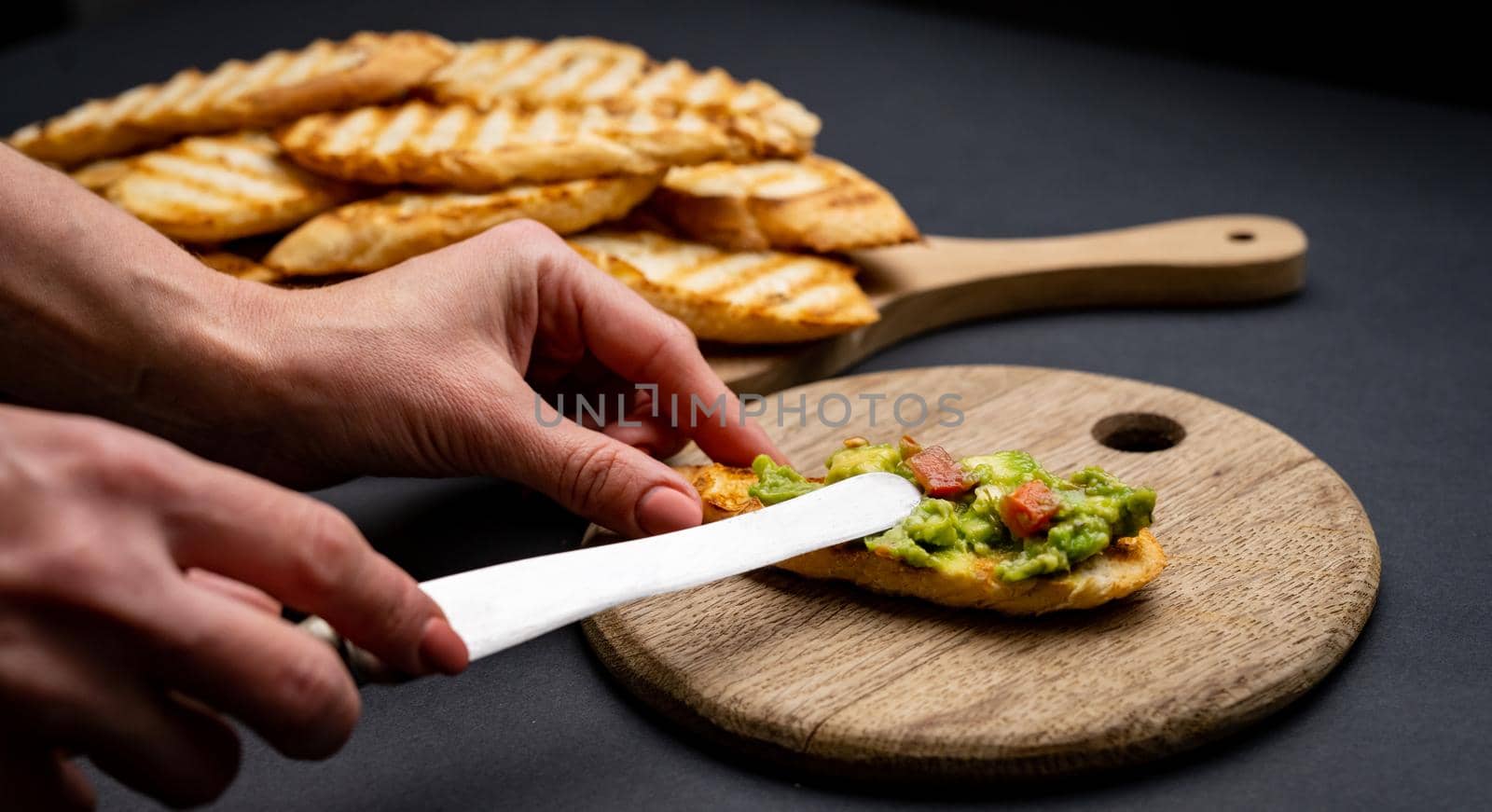 Bruschettas with toast, guacamole and cherry tomatoes on wooden board. Person hands prepare with knife mexican snack slice bread with avocado and vegetables