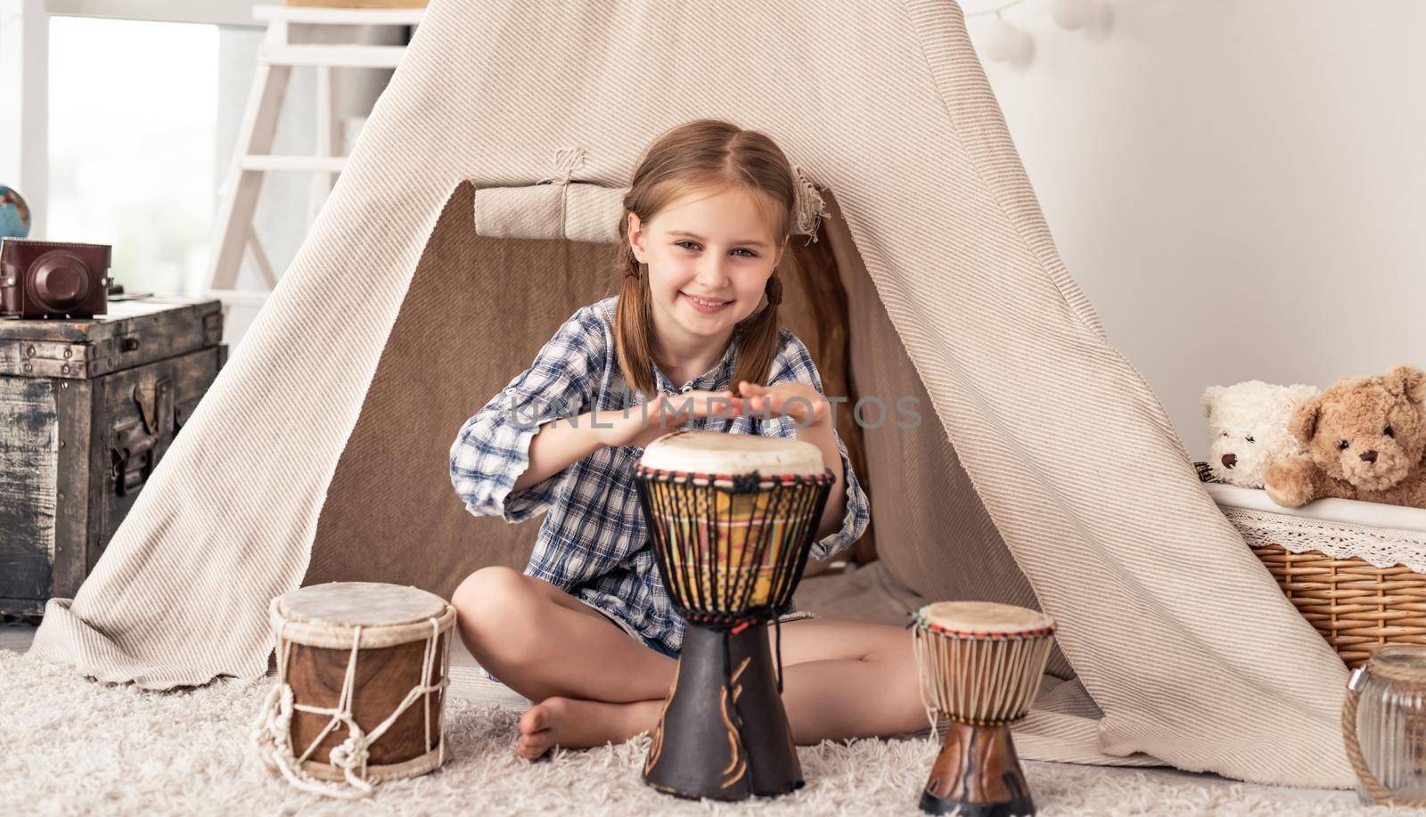 Little girl playing on traditional african djembe drums sitting in wigwam in kids room