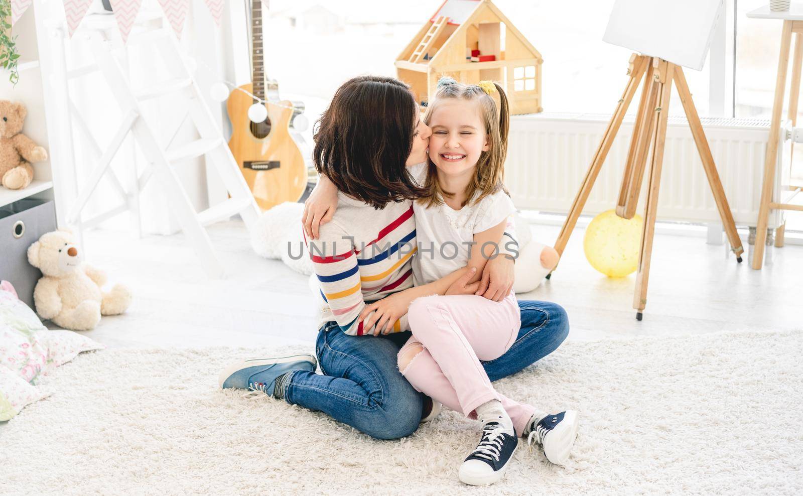 Beautiful mother kissing cute little girl in bright kids room