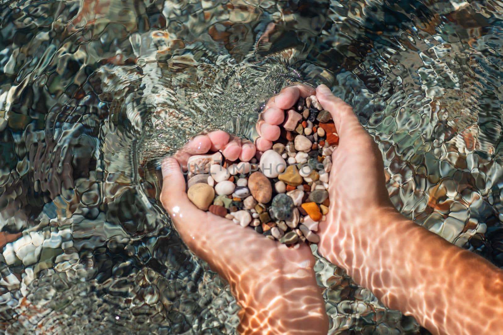 Hands full of colorful pebbles forming heart shape on the sea waves background. Holiday, rest, vacation at the seaside by Len44ik