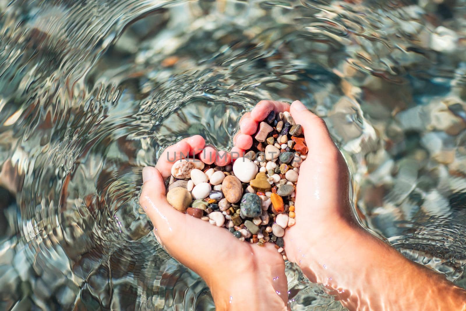 Hands full of colorful pebbles forming heart shape on the sea waves background. Holiday, rest, vacation at the seaside by Len44ik