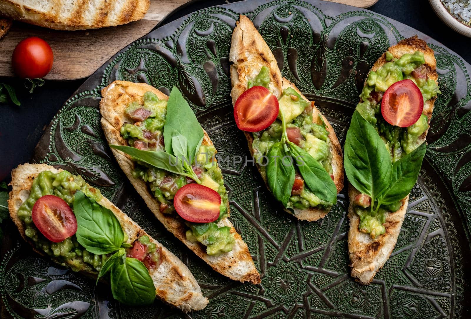 Bruschettas french toasts with guacamole, basil and cherry tomatoes served on beautiful plate with arugula herbs. Mexican snack antipasti with slice bread, mashed avocado and vegetables