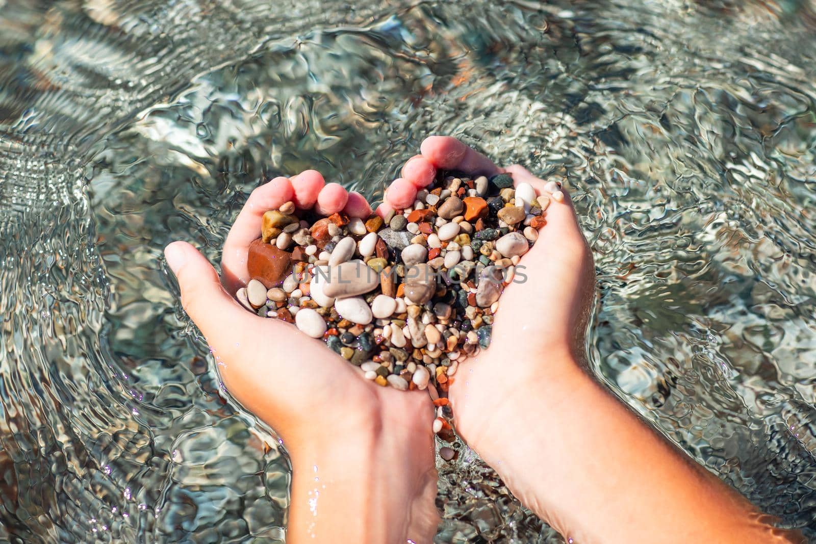 Hands full of colorful pebbles forming heart shape on the sea waves background. Holiday, rest, vacation at the seaside by Len44ik