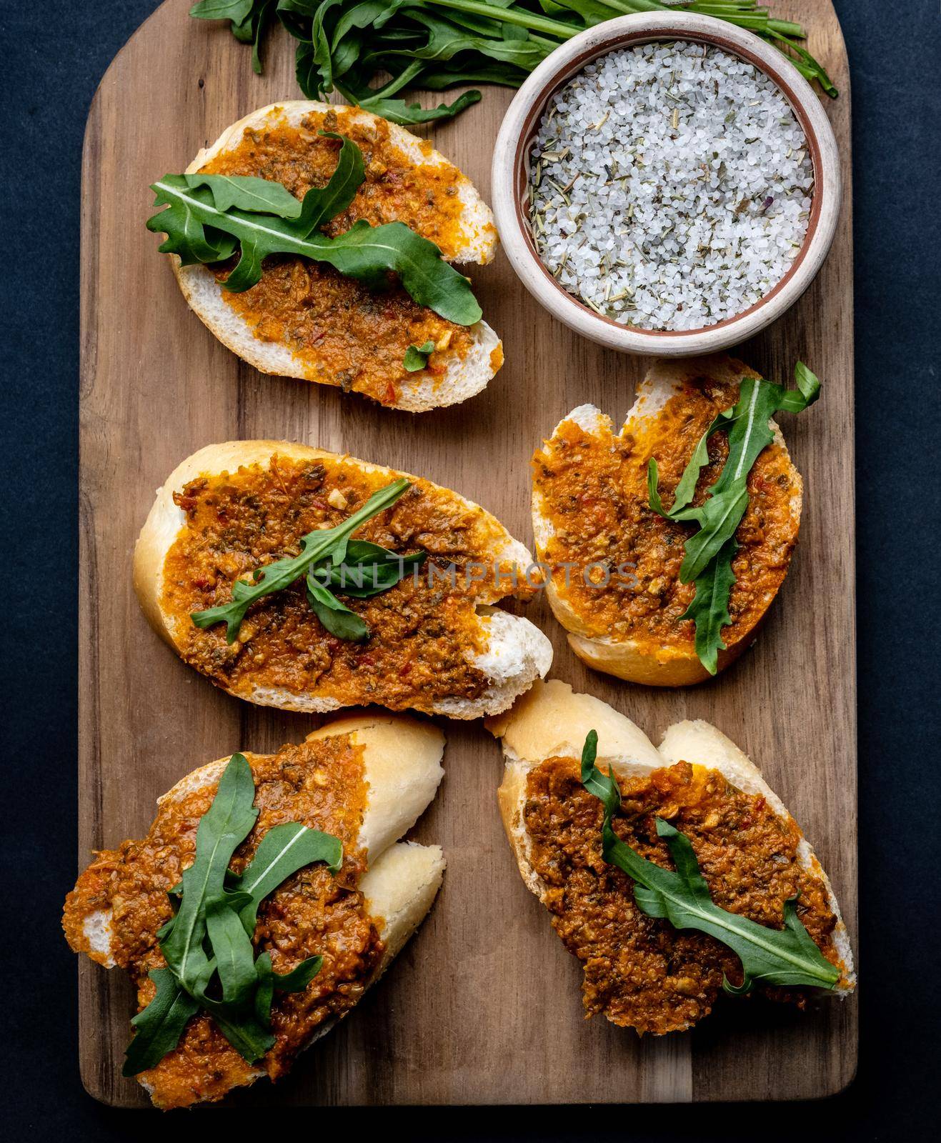 Mediterranean bruschettas with pesto and arugula served on wooden board with salt and herbs. Traditional italian toasted bread with sauce and plants antipasti, closeup view from above