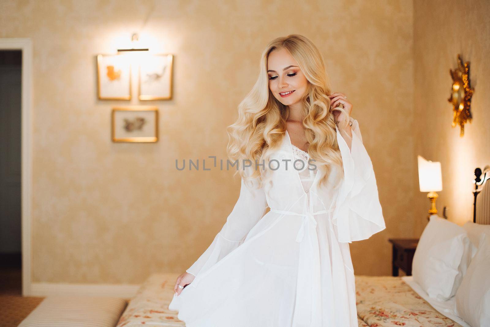 Beautiful and sexy bride wearing in white lace lingerie posing at luxury bedroom interior. Hot blonde haired woman holding dress by hand, sensuality looking at down. Concept of wedding.