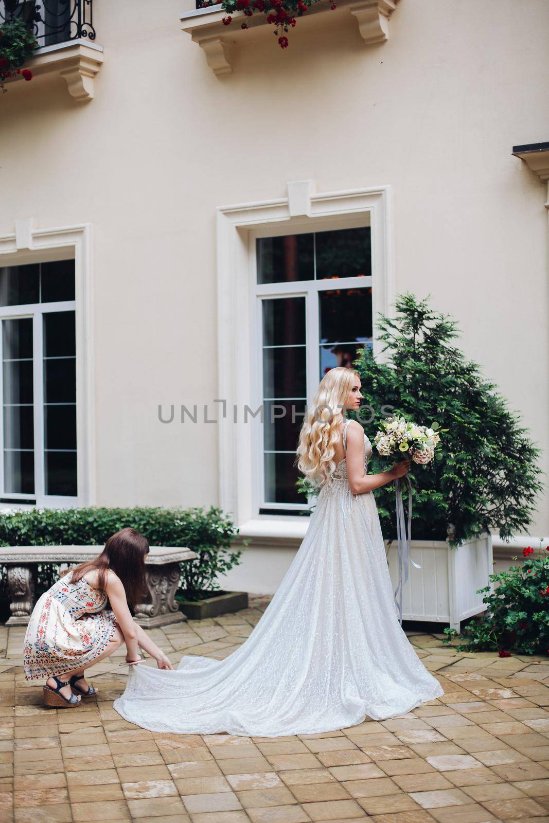 View of gorgeous and sensual bride in perfect, stylish wedding dress posing outdoors, against luxury house with wedding boquet. Beautiful wife waiting for her groom.