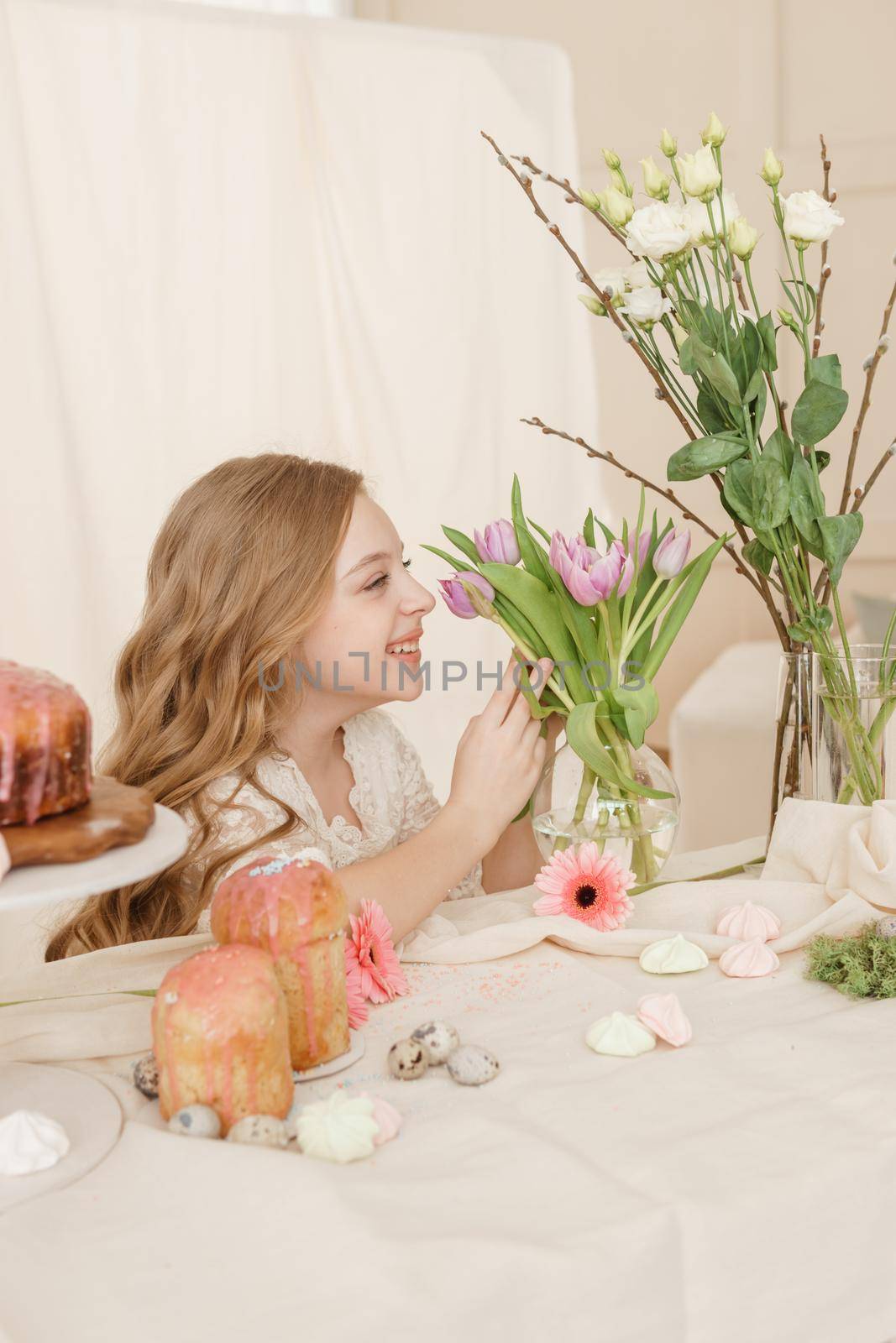 A girl with long hair in a light dress is sitting at the Easter table with cakes, spring flowers and quail eggs. Happy Easter celebration