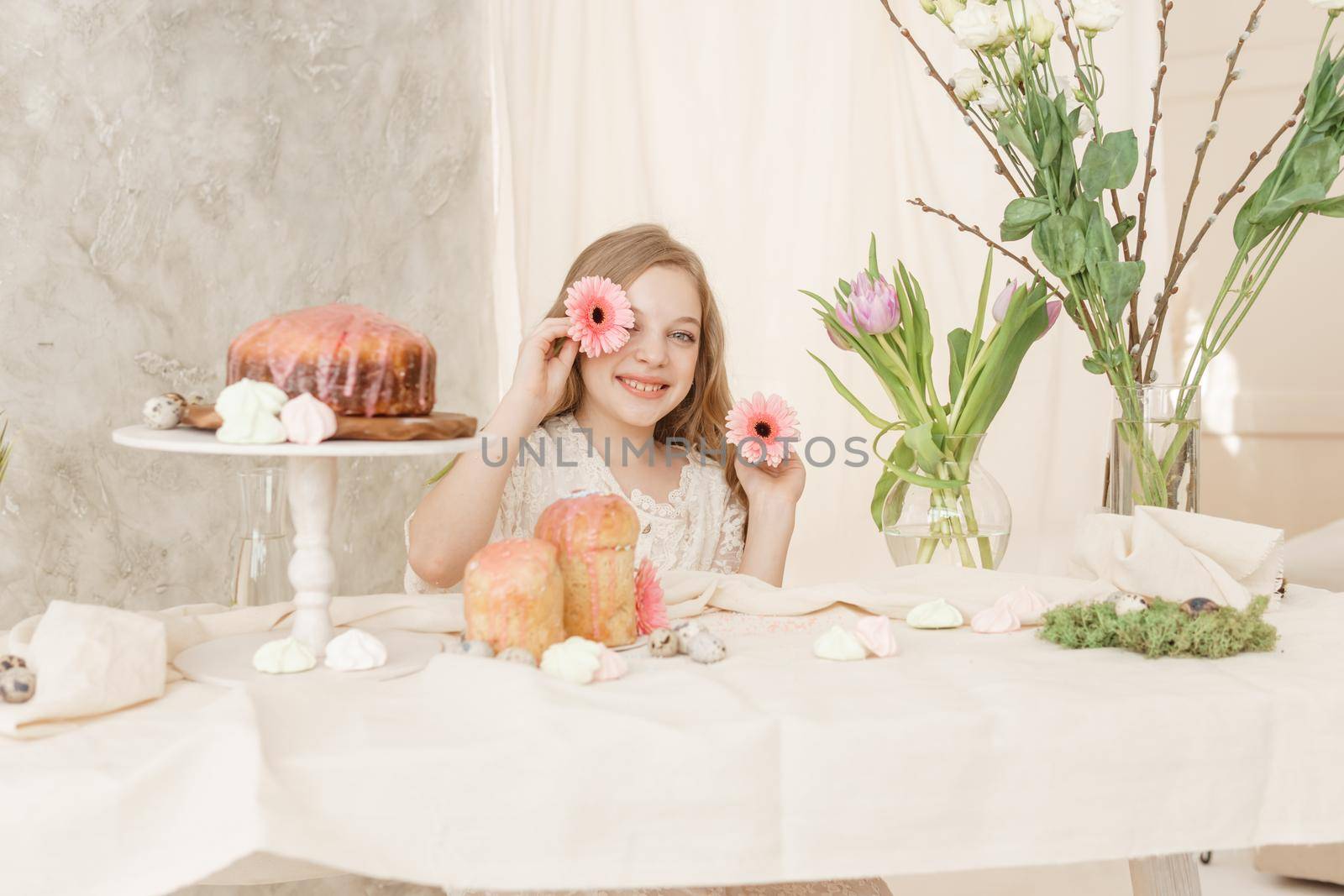 A girl with long hair in a light dress is sitting at the Easter table with cakes, spring flowers and quail eggs. Happy Easter celebration