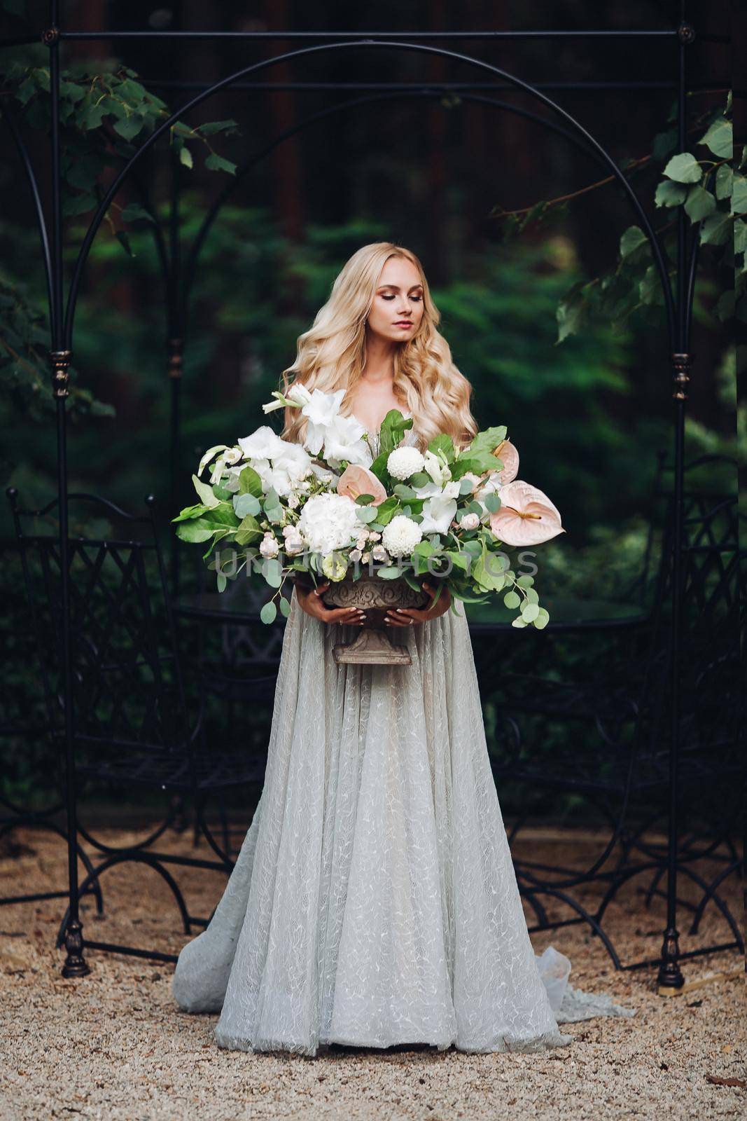 Adorable elegant, sensual young woman looking down and holding wedding flowers boquet. Front view horizontal picture of beautiful blond makeup bride in wedding dress full of love posing on camera.