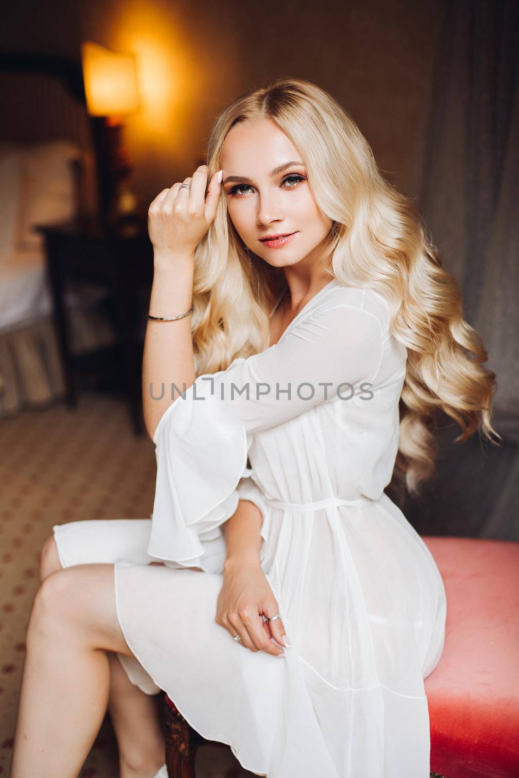 Beautiful and sensuality blondie bride sitting in bedroom, dreaming with closed eyes. Young and sexy woman with long curly hair and perfect make up, wearing in lace lingerie. Morning before wedding.