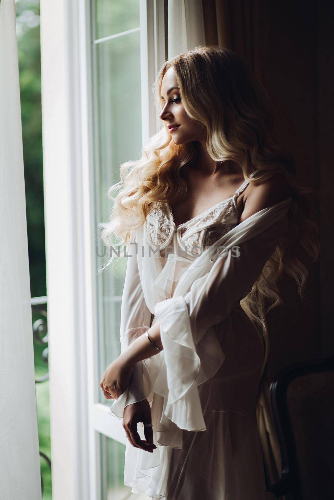 View of gorgeous woman standing near balcony and posing in white lace lingerie. Girl in vintage interior room, looking in big window. Bride at morning.