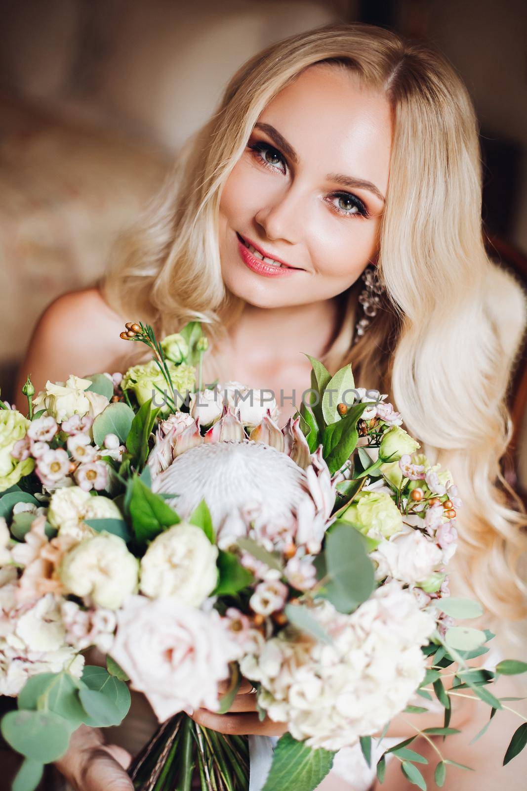 Portrait of beautiful blonde with attractive body, wearing in elegant white lace lingerie looking downn on flowers. Pretty bride holding wedding bouquet in one hand and posing with closed eyes.