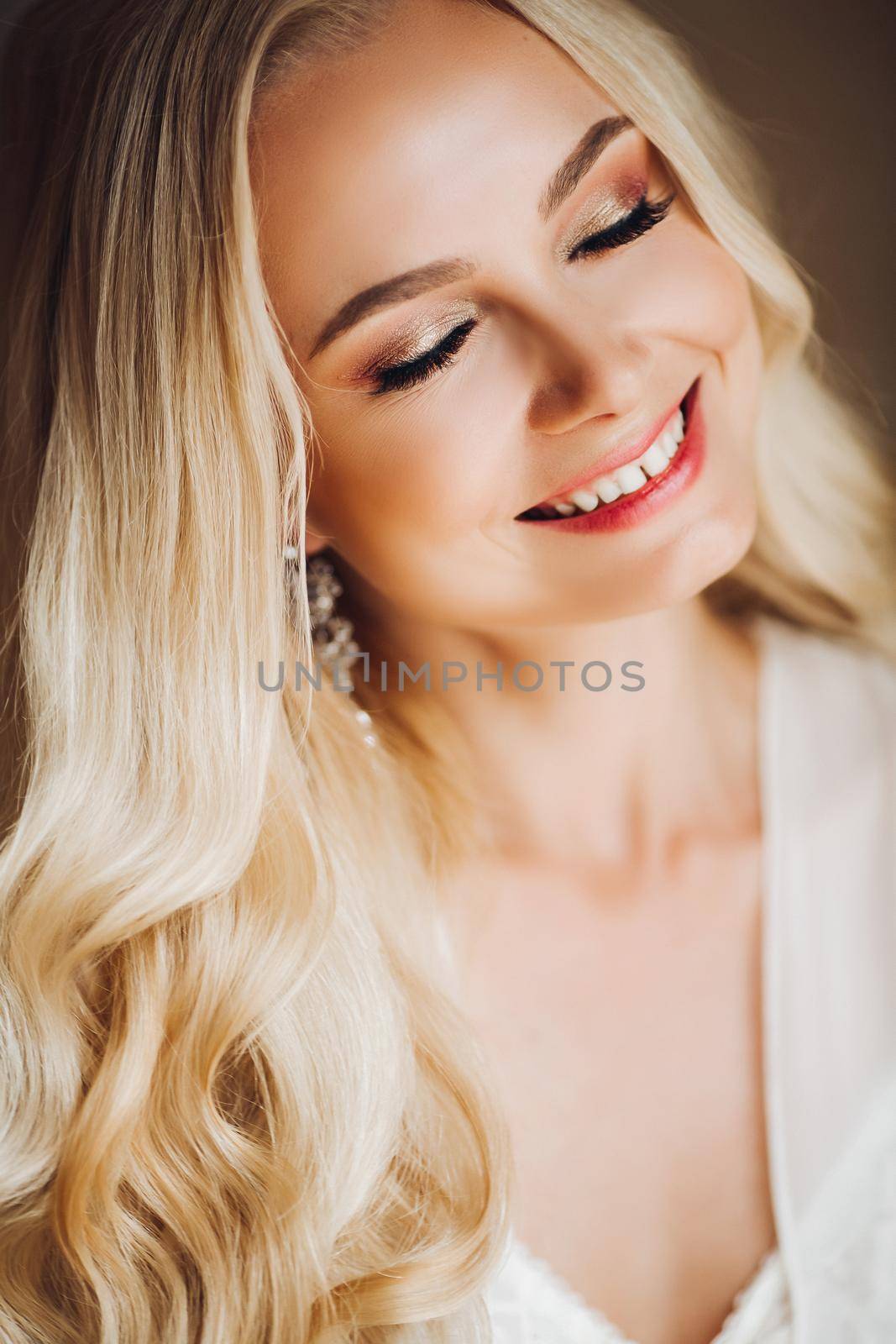 Beautiful and sensuality blondie bride sitting in bedroom, dreaming with closed eyes. Young and sexy woman with long curly hair and perfect make up, wearing in lace lingerie. Morning before wedding.