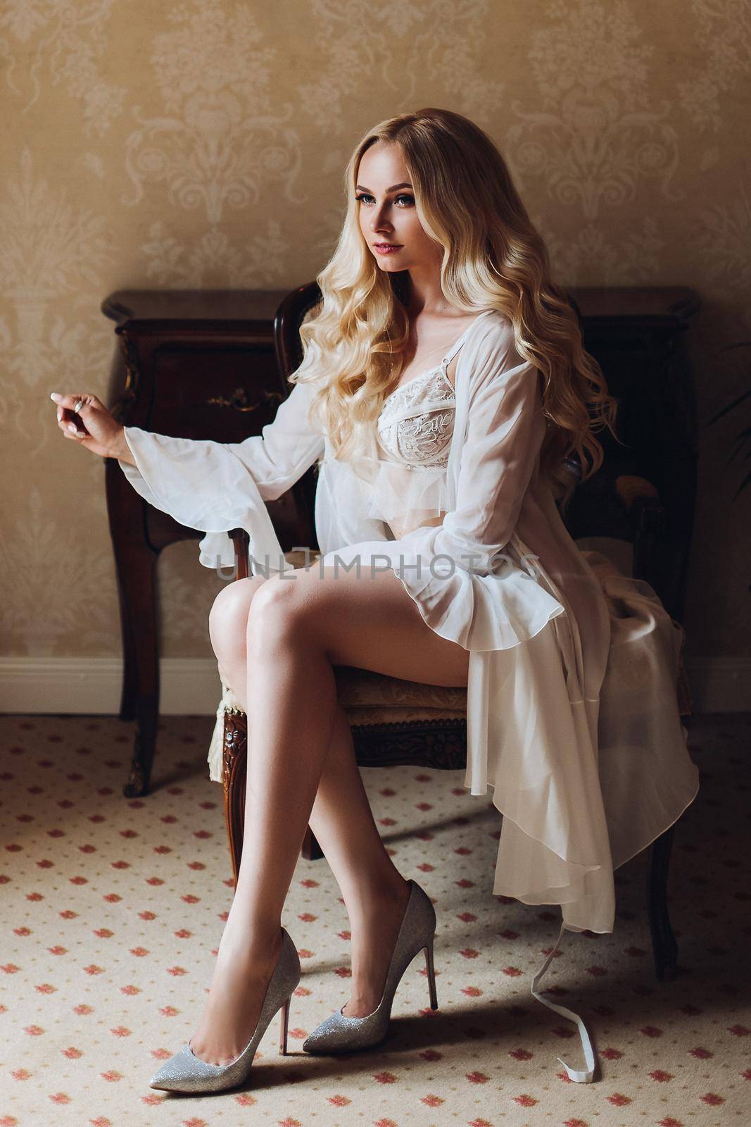 Gorgeous and beautiful blondie bride with long wavy hair, sitting on chair in luxury interior, looking at camera. Seductive woman wearing in white lace lingerie in morning. Wedding concept.