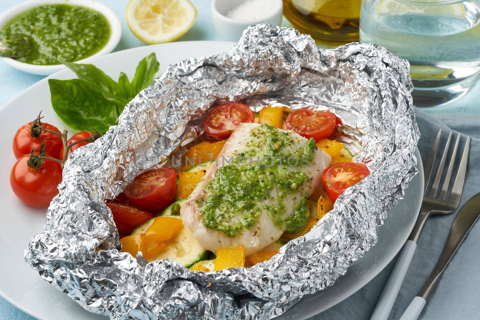 Foil pack dinner with white fish. Oven baked fillet of cod, pike perch with vegetables and pesto sauce. Healthy diet food, keto diet, Mediterranean cuisine. Blue background. Side view