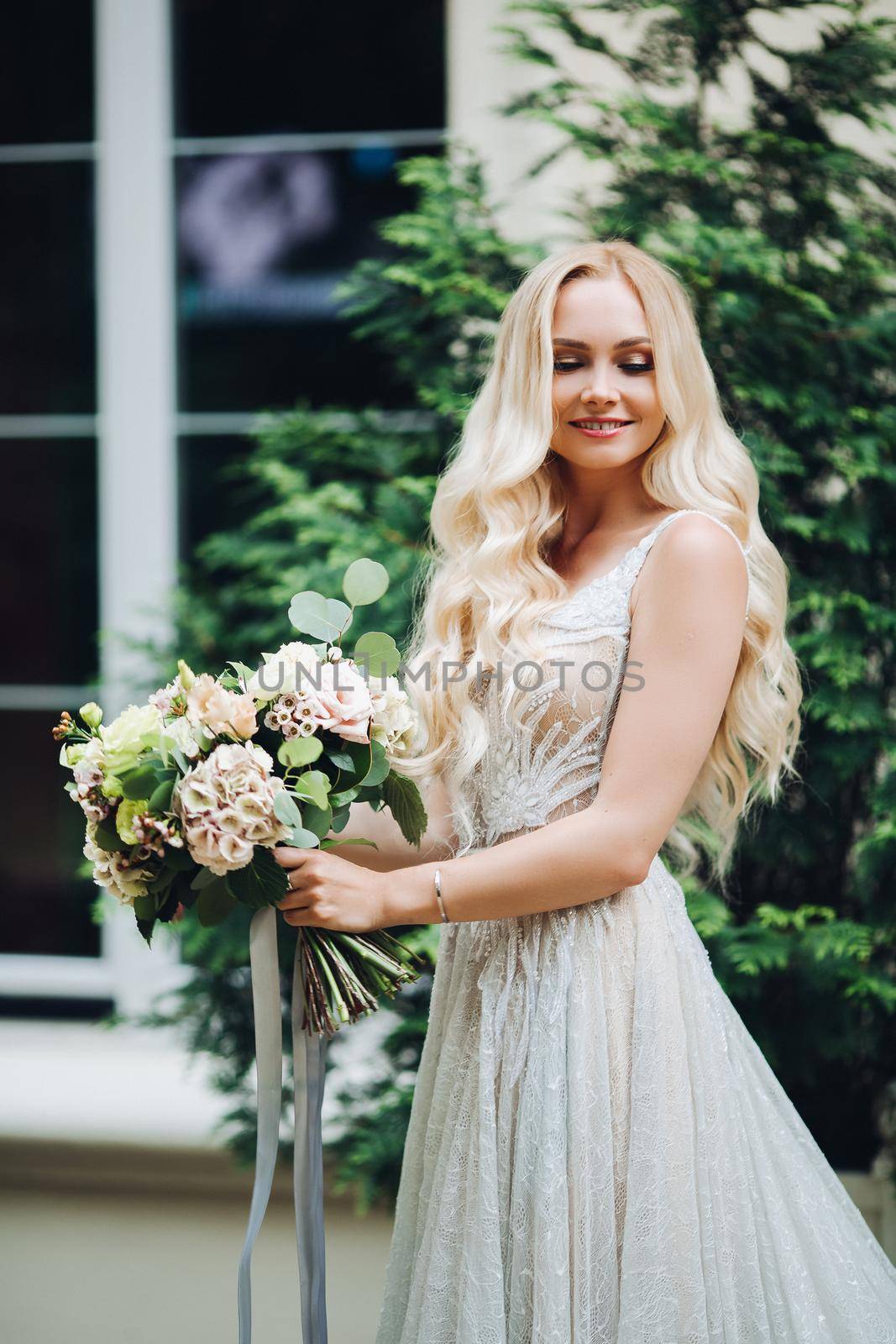 Front view of elegant and sensuality blondie bride looking at camera and smiling, holding wedding boquet. Beautiful female with makeup, wearing in wedding dress, posing outdoors.