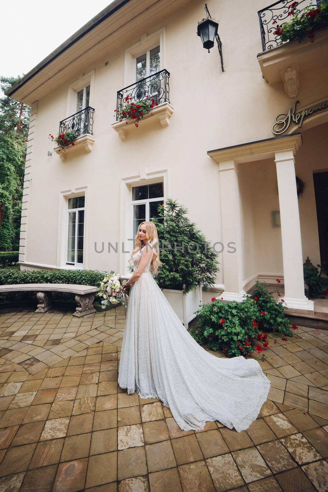 View of gorgeous and sensual bride in perfect, stylish wedding dress posing outdoors, against luxury house with wedding boquet. Beautiful wife waiting for her groom.