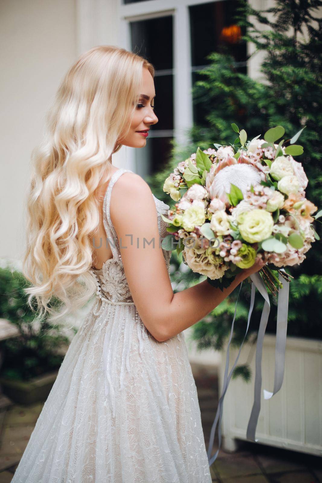 View of gorgeous and sensual bride in perfect, stylish wedding dress posing outdoors, against luxury house with wedding boquet. Beautiful wife waiting for her groom.