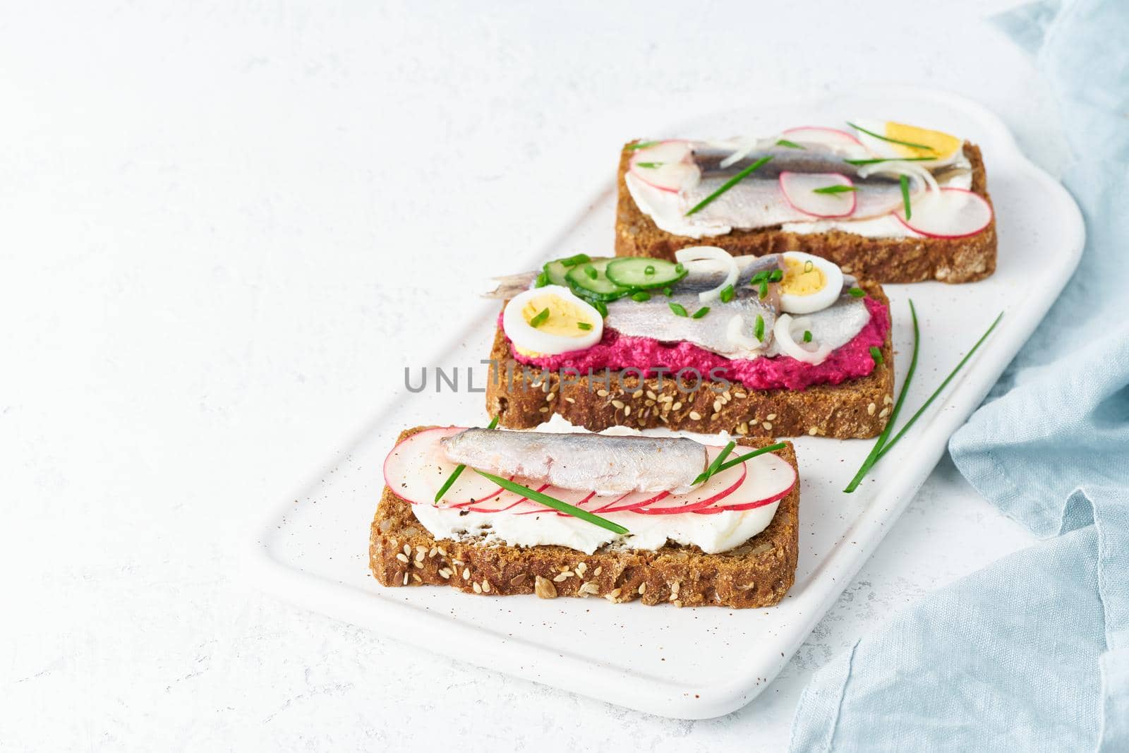 Savory smorrebrod, set of traditional Danish sandwiches. Black rye bread with anchovy, beetroot, radish, eggs, cream cheese on grey plate on a white stone table, side view, copy space