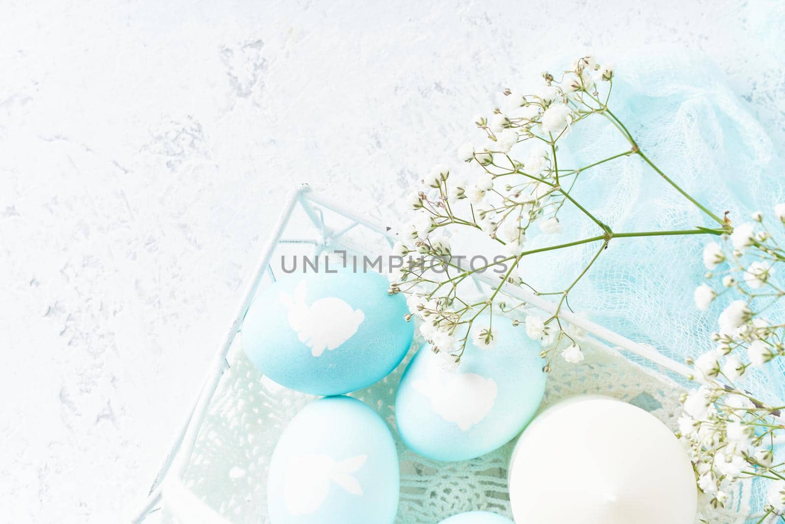 Easter. Holiday. Light white background, gentle pastel colors. Blue eggs with image of rabbit in basket. Flowers in background, top view, copy space,