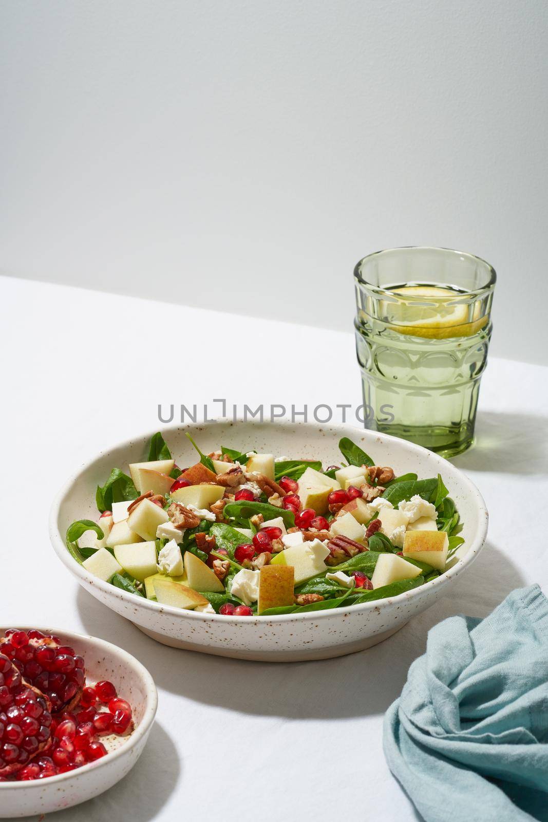 Fruits salad with nuts, balanced food, clean eating. Spinach with apples, pecans and feta, garnished with pomegranate seeds in bowl on table with white tablecloth. Hard light, shadows, vertical