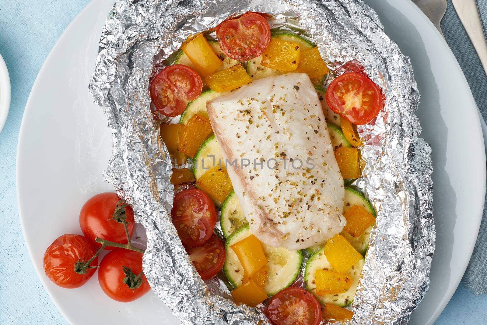 Foil pack dinner with white fish. Oven baked fillet of cod, pike perch with vegetables and pesto sauce. Healthy diet food, keto diet, Mediterranean cuisine. Blue background. Top view, close up