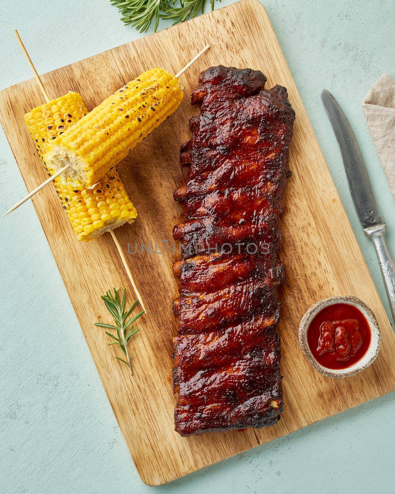 Barbecue pork ribs. Slow cooking recipe. Whole pickled roasted pork meat with red sauce and corn on cutting board, vertical, top view. Turquoise background