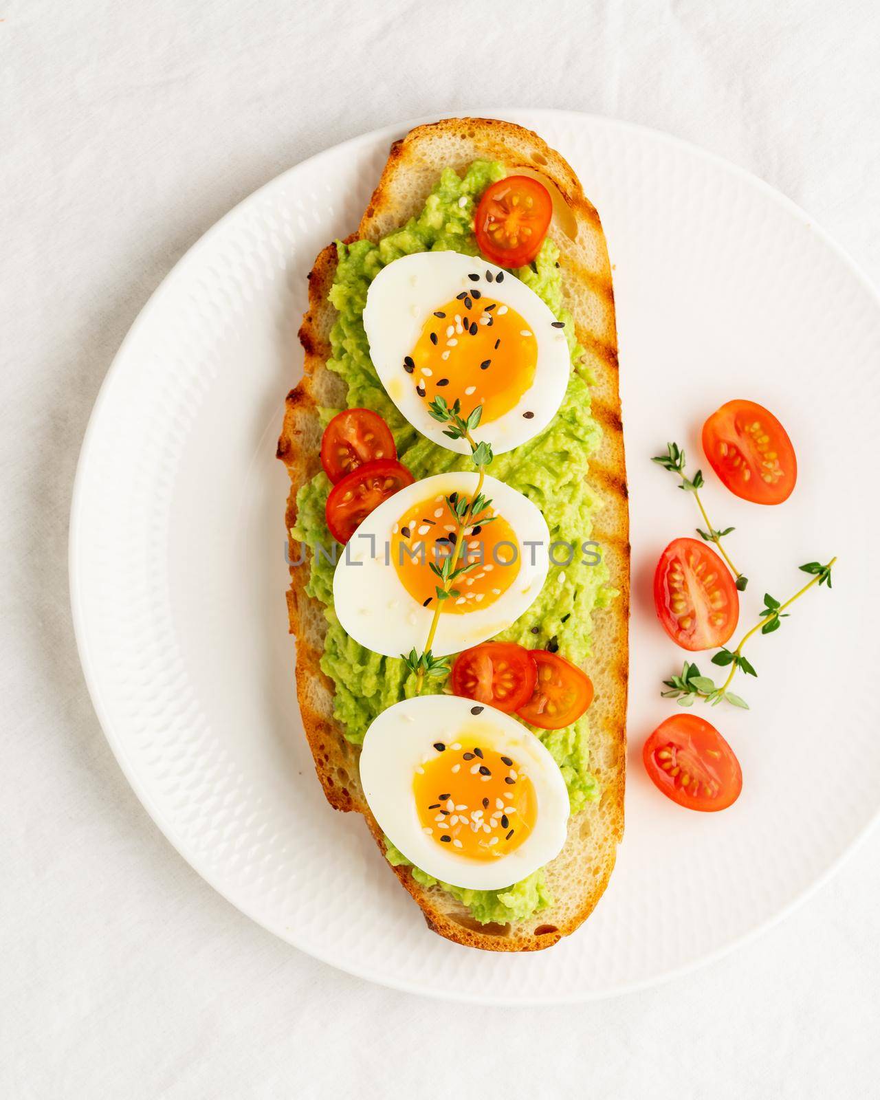 Avocado toast with toasted bread soft-boiled eggs with yellow yolk and tomatoes with herbs on white plate on light tablecloth